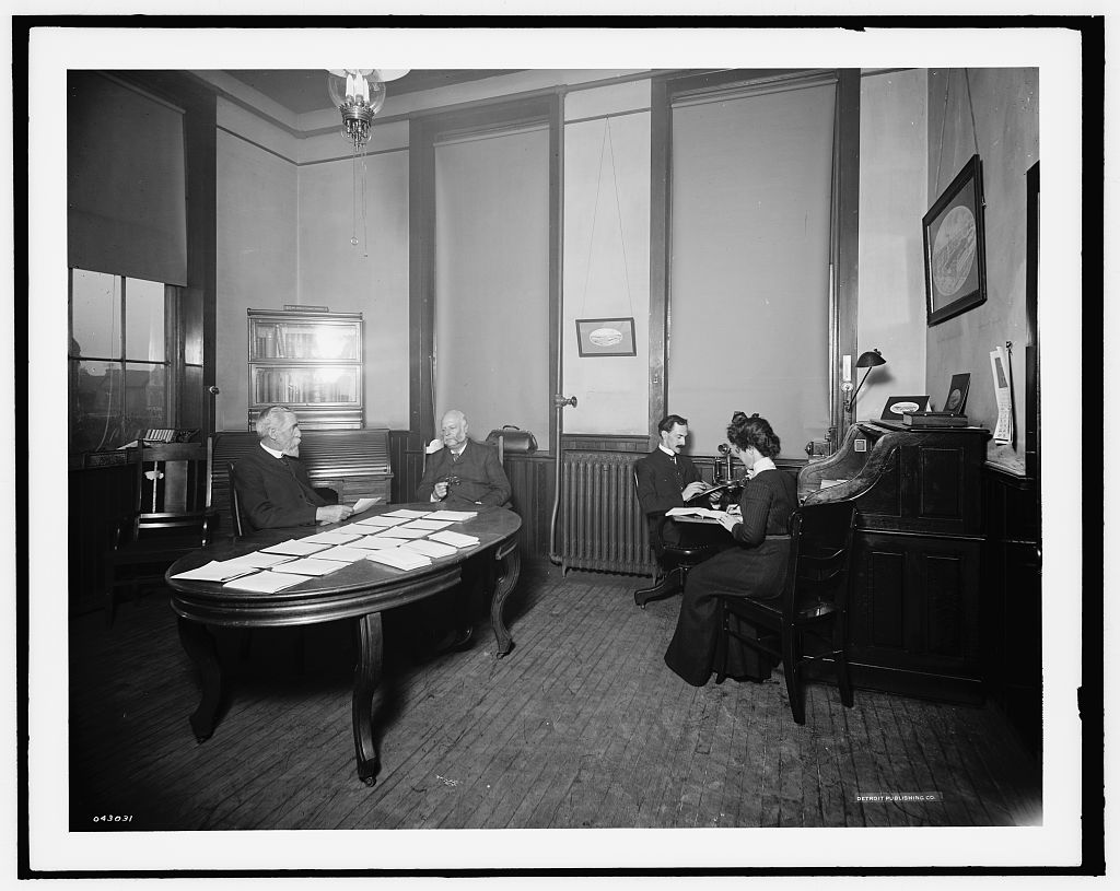 An early twentieth-century manufacturing office with people gathered at a desk and drafting station.