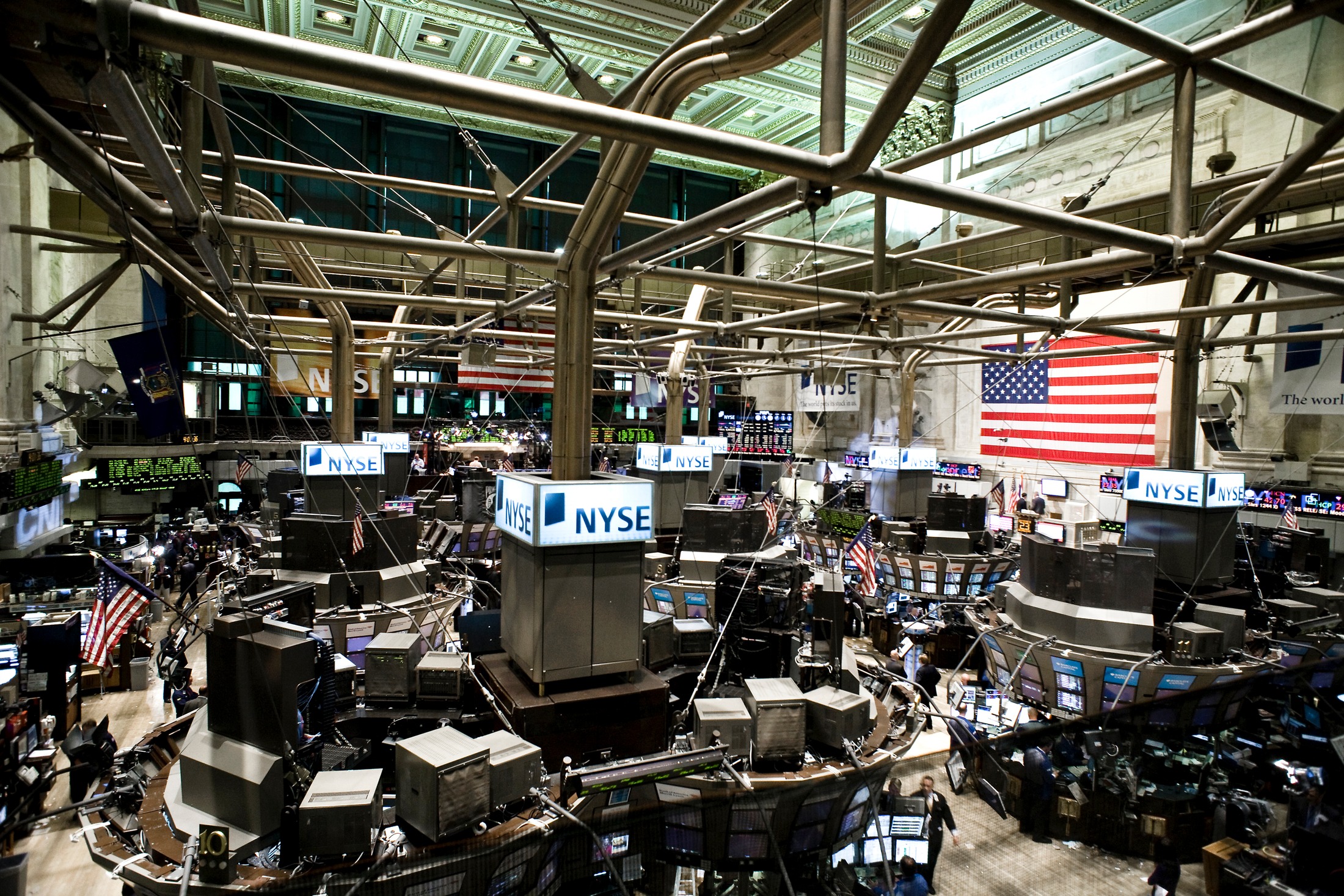 A wide view of the New York Stock Exchange trading floor, with trading posts, screens, overhead walkways, and an American flag.